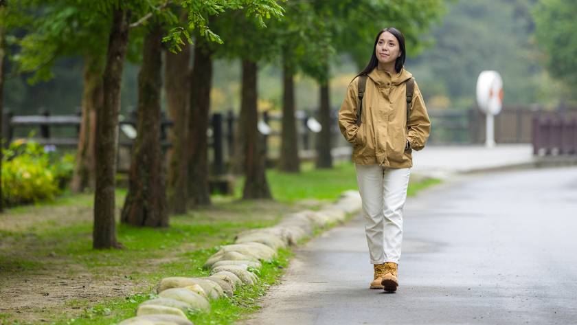 Woman walking through serene park path lined with trees and stones, wearing a tan jacket and boots