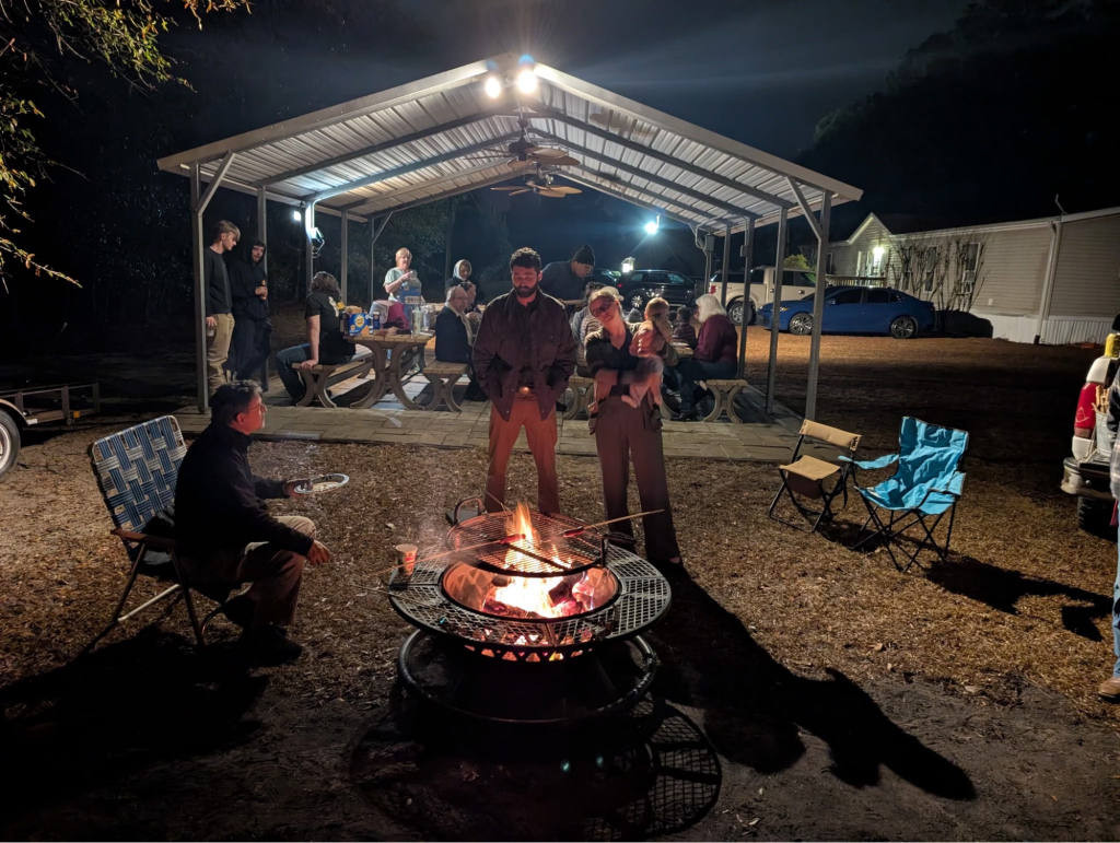 People gathered around a bonfire at night with picnic tables under a shelter, enjoying a cozy outdoor gathering