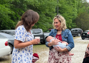 Woman holding baby and chatting outdoors with another woman in floral dress next to parked cars