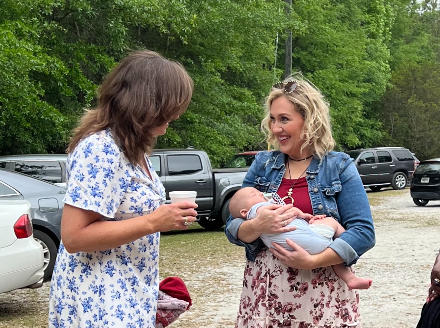 Woman holding baby and chatting outdoors with another woman in floral dress next to parked cars