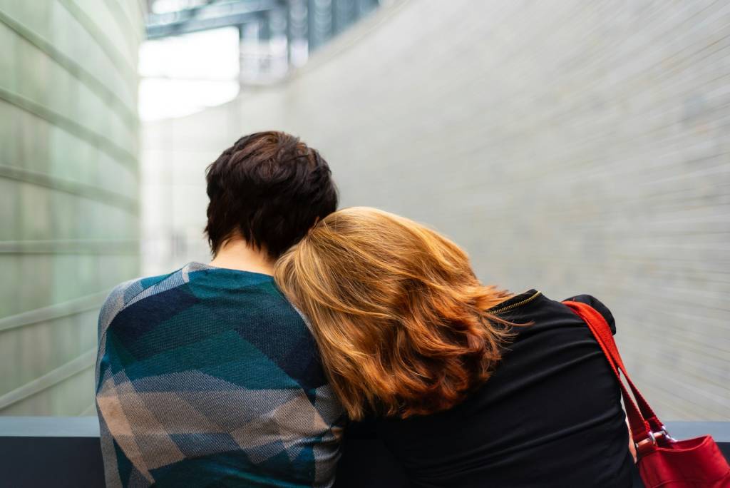 Two people sitting closely in a modern space, one resting their head on the other's shoulder, conveying comfort and support