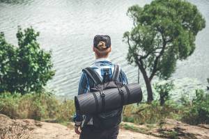 Hiker with backpack and camping mat overlooking serene lake surrounded by trees