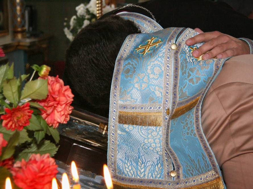 Priest in ornate blue vestment performing a religious ritual, surrounded by flowers and burning candles