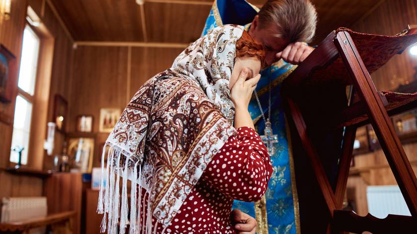 Woman in headscarf confesses to priest in traditional robes inside a wooden church setting