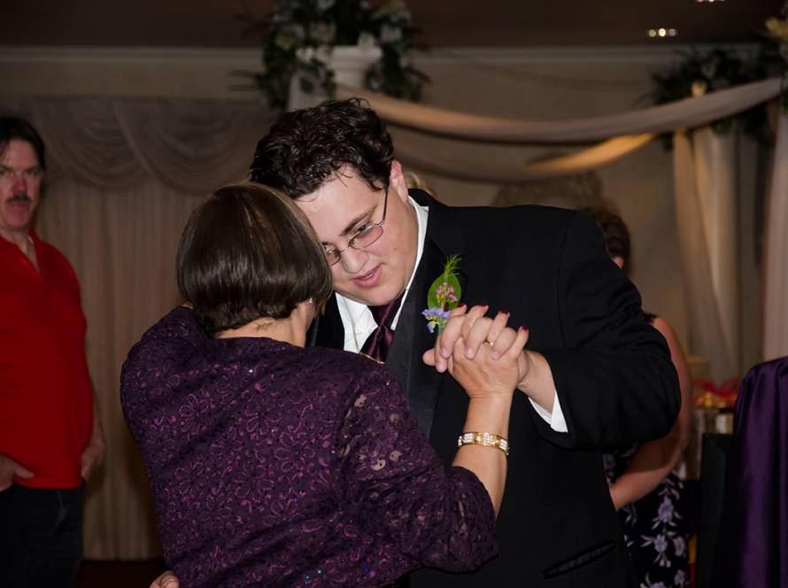 Mother-son dance at a wedding reception, guests in the background, elegantly dressed in formal attire