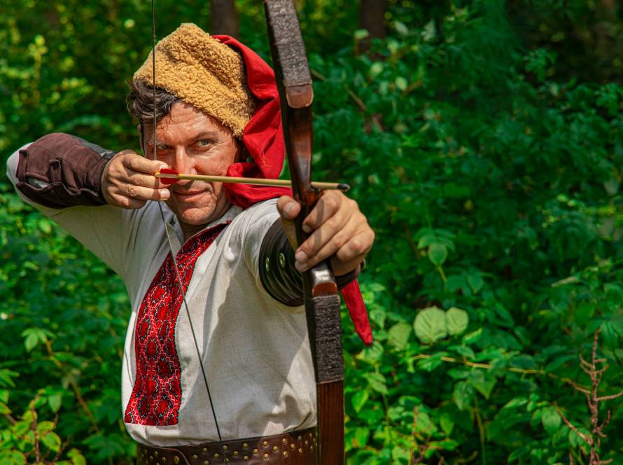 Medieval archer in traditional attire aiming a bow in a lush forest setting