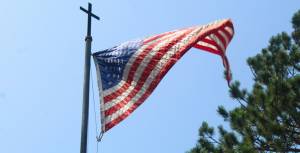 American flag waves on a tall pole with cross top against clear blue sky, trees in the background.