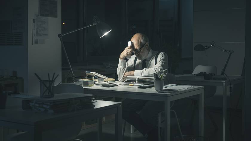 Man working late in dimly lit office, using a phone, surrounded by papers and desk lamp illuminating workspace