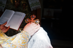 Priest's hand holding ornate cross and open book during religious ceremony in ornate robe