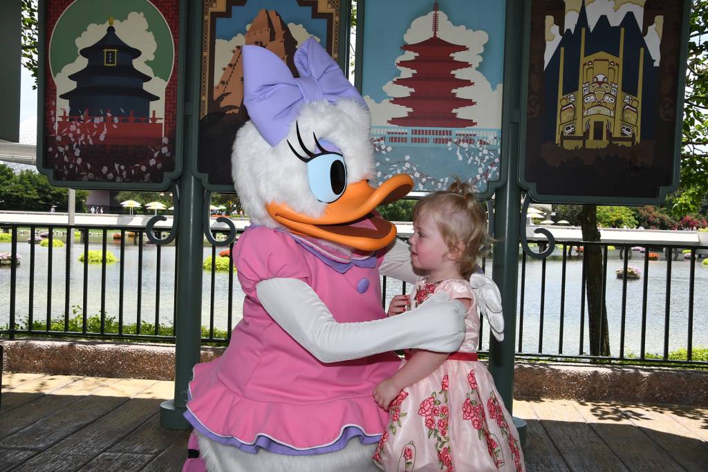 Child in floral dress meets a duck character in a pink outfit by lakeside with themed posters in background
