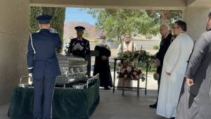 Military funeral ceremony with honor guard saluting and attendees in formal attire near casket and floral arrangements