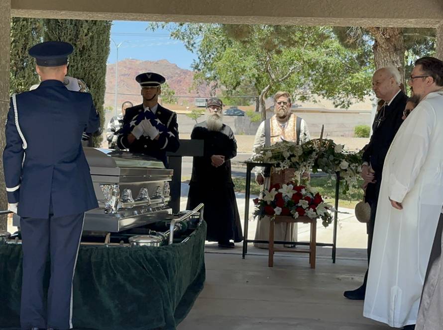 Military funeral ceremony with honor guard saluting and attendees in formal attire near casket and floral arrangements