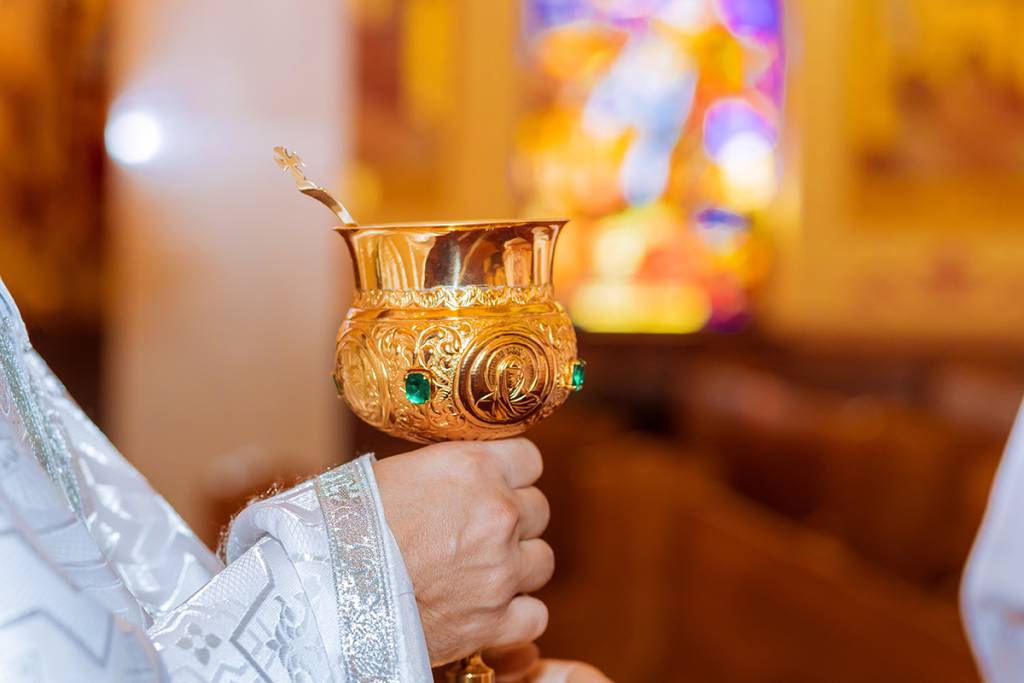 Golden chalice adorned with emeralds held by priest during church ceremony with stained glass background