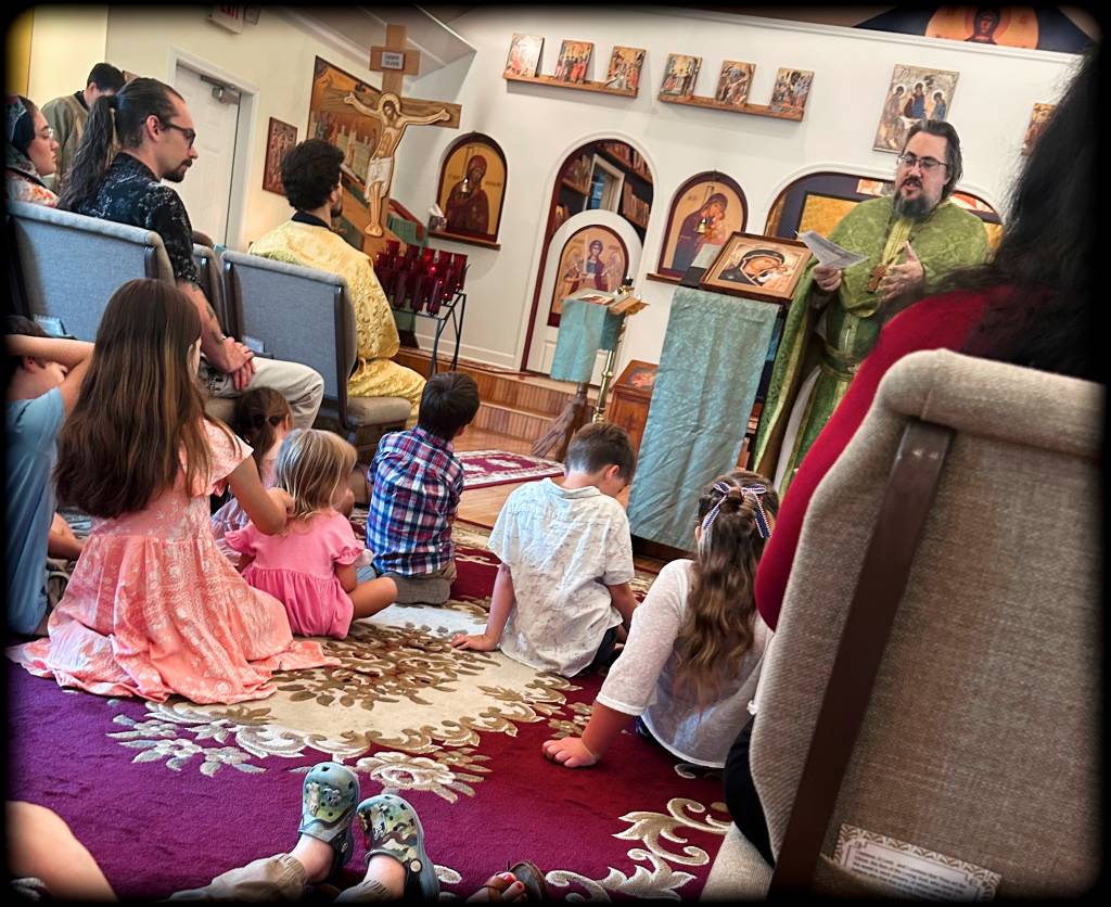 Church service with children sitting on a rug, listening to a speaker in green robes, surrounded by religious icons