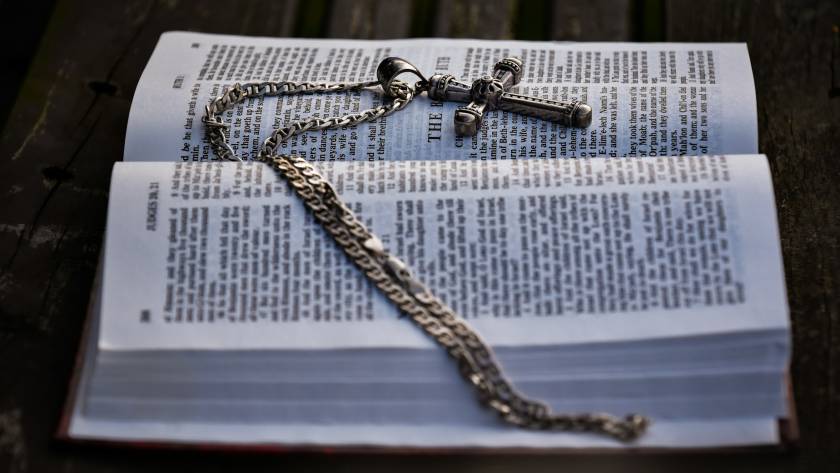 Open Bible with ornate silver cross necklace lying on its pages outdoors on wooden surface