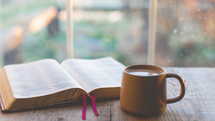 Open book and coffee mug on wooden table by window, creating a cozy reading atmosphere