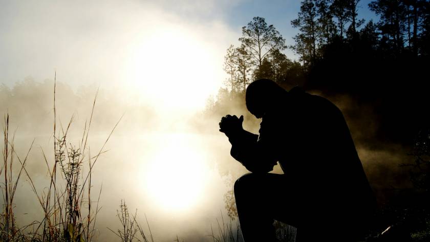 Silhouette of a person kneeling in prayer by a misty lake at sunrise surrounded by trees