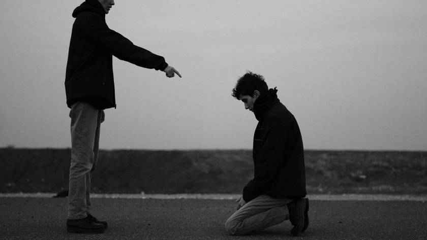 Person pointing aggressively at someone kneeling on the ground in a dark, moody scene