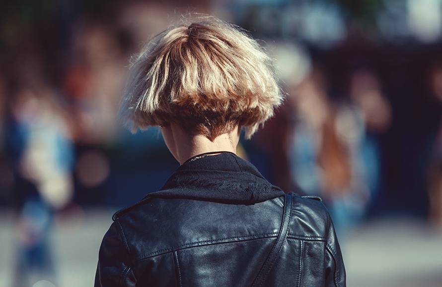 Person with short blonde hair in black leather jacket standing outdoors in a blurred urban background