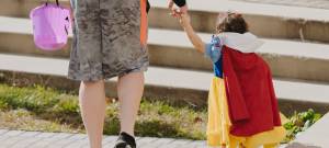 Child in princess costume trick-or-treating, holding adult's hand, carrying purple candy bucket